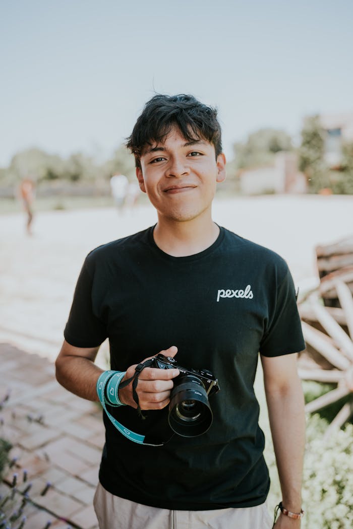 services-04 A smiling young man with a camera poses casually outdoors, ideal for lifestyle and photography themes.